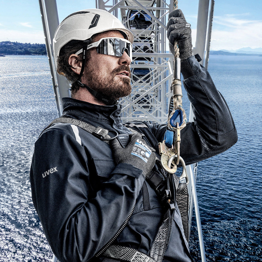 A man in diving or offshore work gear stands on a metal tower over blue water, wearing a white helmet, mirrored sunglasses, black gloves, and a black Uvex jacket with harness straps and carabiners.